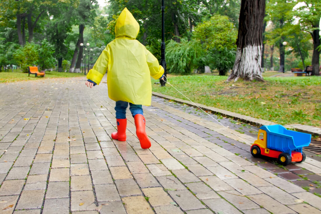 Cute little boy in yellow raincoat running with toy truck under rain at park