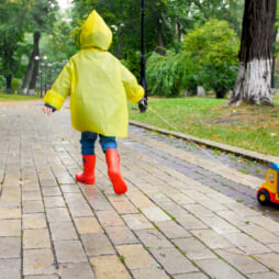 Cute little boy in yellow raincoat running with toy truck under rain at park